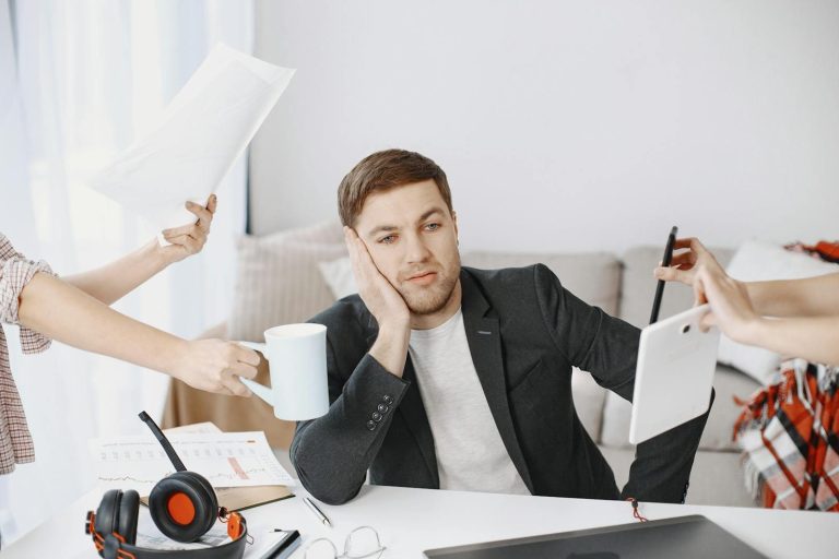 Overwhelmed man at desk surrounded by hands holding work items.